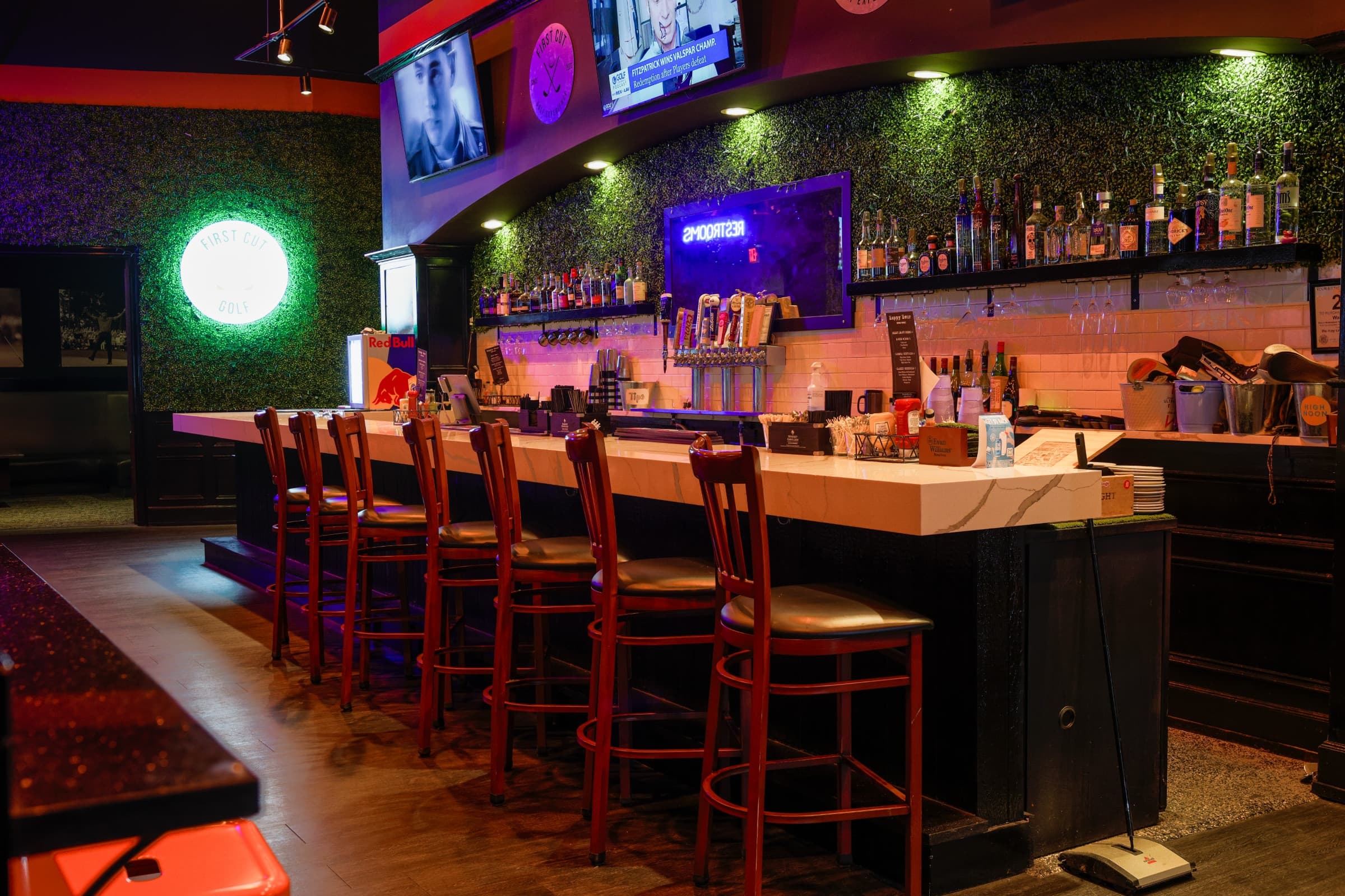 The bar at First Cut Golf — marble bar top, full bottle wall on backlit shelves, row of stools, and the illuminated FIRST CUT GOLF mark on the wall.