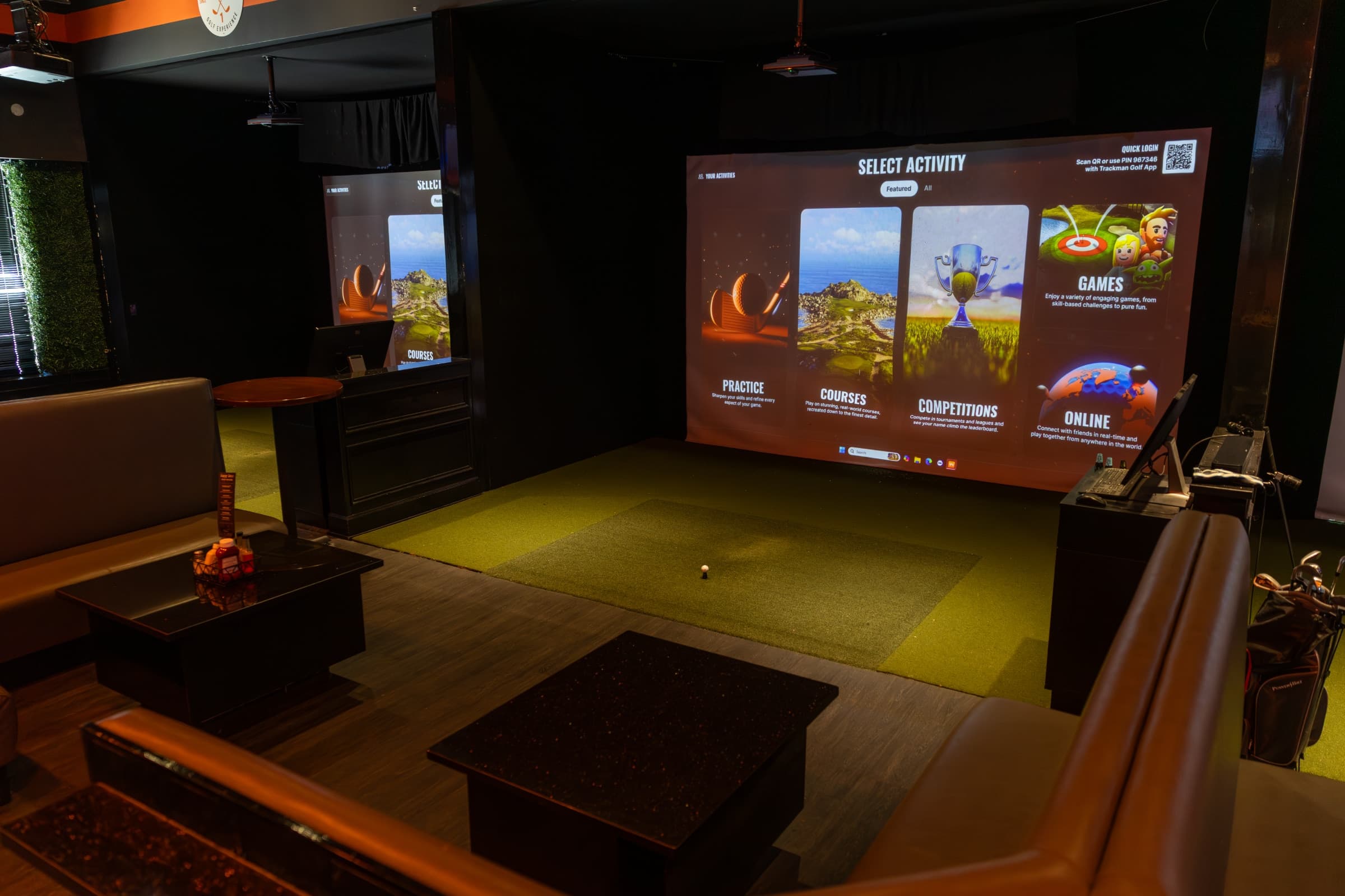 A simulator bay at First Cut Golf — leather L-shaped booth wrapped around a low table, with the TrackMan IO screen showing the activity menu and a turf putting strip leading to the screen.